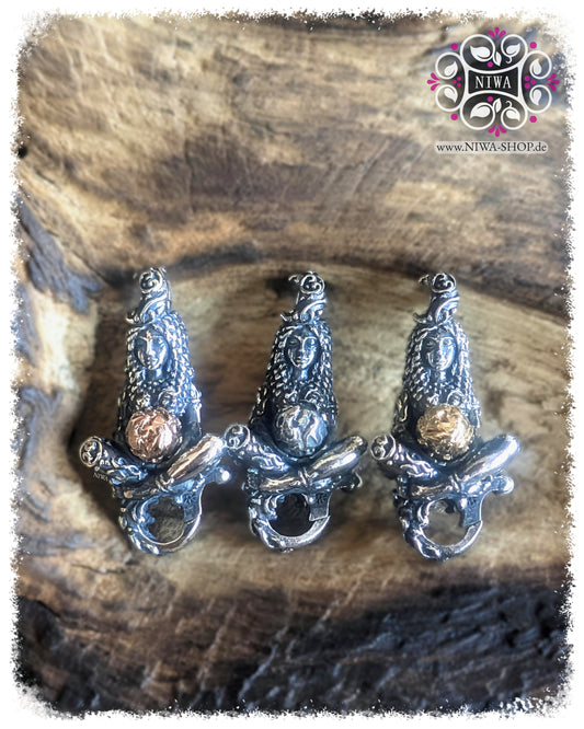 GAIA MOTHER of LIFE Lock by MAMA MALA in three versions on weathered wood. In the middle the silver version with a silver globe, on the left the two-tone version with a rose gold ball, on the right the two-tone version with a yellow gold ball. The seated female figure carries the world in her hands and shows organic leaves, tendrils and root structures.