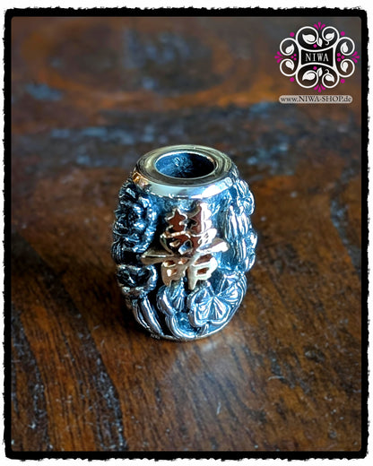 CHINESE DOUBLE HAPPINESS BarrelCharm aus 925 STERLINGSILBER mit 18K ROSÉGOLD Schriftzeichen und LOTUSBLÜTEN-Relief, frontal auf dunkler Holzoberfläche vor NIWA Logo.