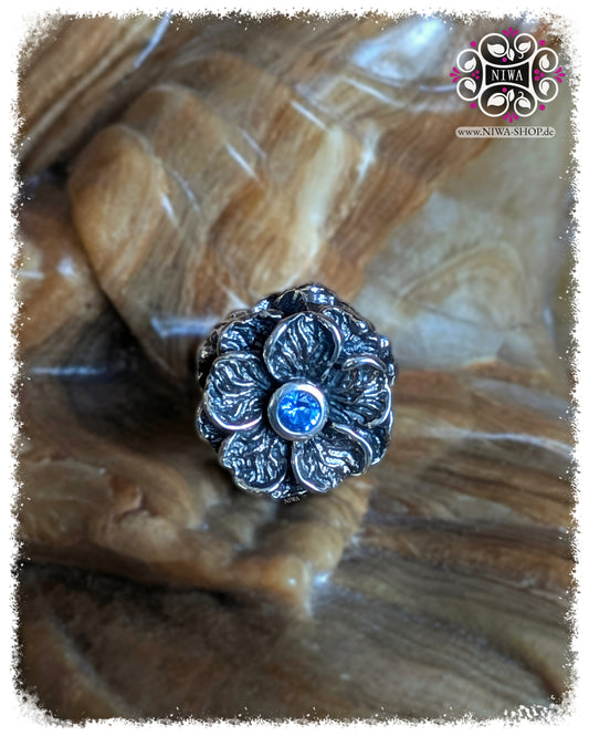 FORGET ME NOT Charm, 925 SterlingSilber, blauer Quarz. Hauptmotiv in Frontalansicht: große Vergissmeinnicht-Blüte mit facettiertem blauem Quarz in der Mitte, plastisch ausgearbeitete Blütenblätter mit dunkleren Vertiefungen, auf Steinuntergrund.
