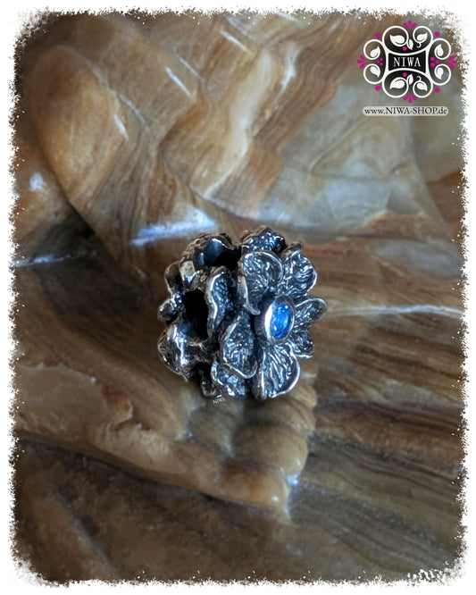 FORGET ME NOT Charm, 925 SterlingSilber, blauer Quarz. Schrägansicht der Blüte mit sichtbarer Tiefe der Blütenblätter und mittigem blauem Quarz, markante, oxidierte Details, auf Steinuntergrund.